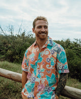 A man with short hair and a beard, wearing the Golden Breed Batik Shirt | Rose, stands smiling outdoors by a wooden fence, with greenery and a cloudy sky in the background.
