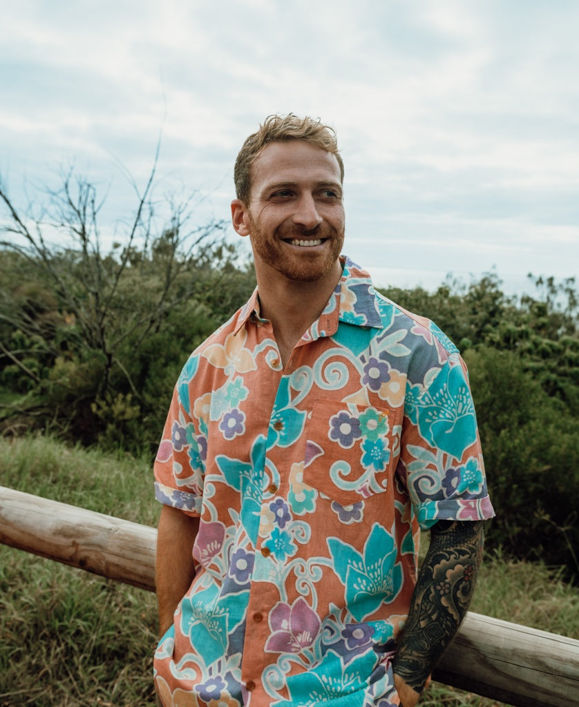 A man with short hair and a beard, wearing the Golden Breed Batik Shirt | Rose, stands smiling outdoors by a wooden fence, with greenery and a cloudy sky in the background.