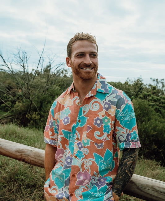 A man with short hair and a beard, wearing the Golden Breed Batik Shirt | Rose, stands smiling outdoors by a wooden fence, with greenery and a cloudy sky in the background.