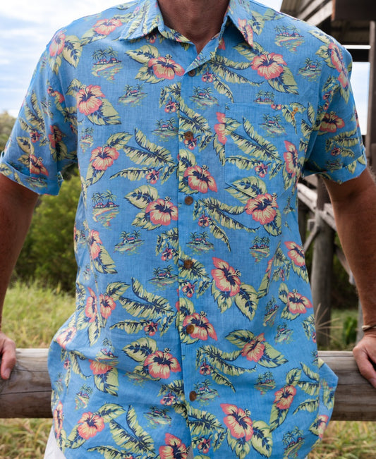 A person wears the Golden Breed Howe Shirt | Azure Blueโan oversized Hawaiian shirt with a blue tropical floral patternโwhile leaning on a wooden rail outdoors.