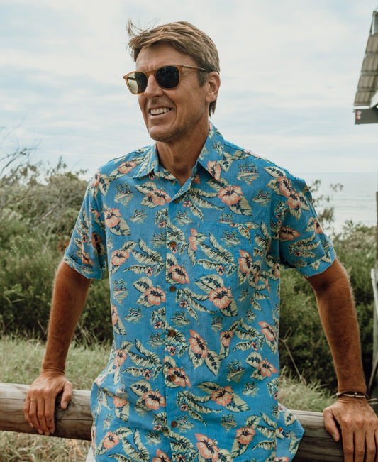 A man wearing the Howe Shirt in Azure Blue by Golden Breed, an oversized-fit Hawaiian shirt crafted from a soft cotton/linen blend, stands outdoors leaning on a wooden fence with greenery and sky in the background.