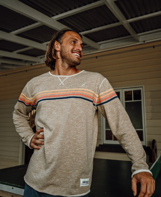 A man with long hair stands indoors, smiling, wearing the Golden Breed Long Island Rag Top in Coffee Marle, featuring colorful horizontal stripes for a retro style look.