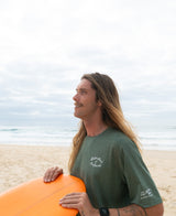 Wearing the Golden Breed Nemo S/S Rash Shirt in Green Marle, a person with long hair stands on the beach holding an orange surfboard, gazing out at the ocean.