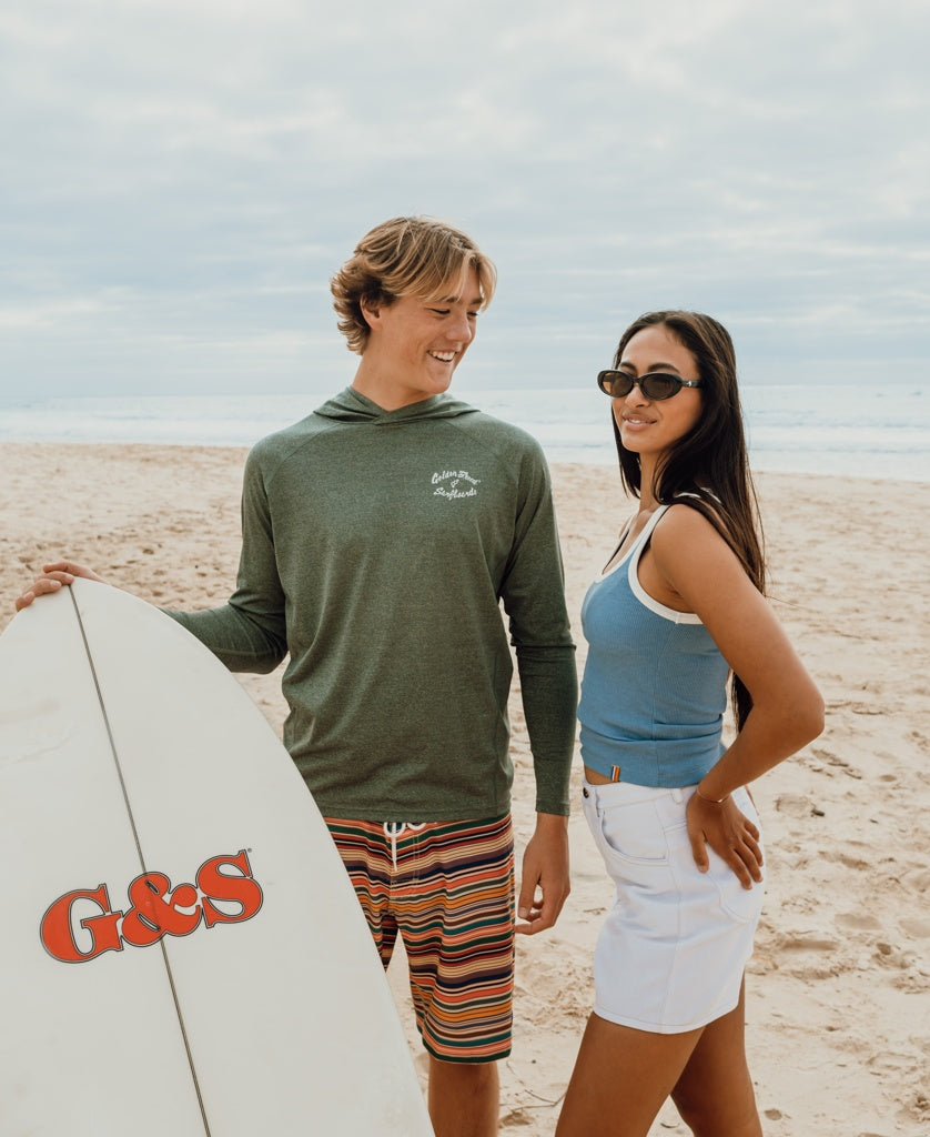 Two people stand on a sandy beach, one holding a surfboard labeled “G&S.” Both wear Golden Breed Rapper Hoodie Rash Shirts in Green Marle, offering 50+ UPF protection. The ocean extends behind them under a cloudy sky.