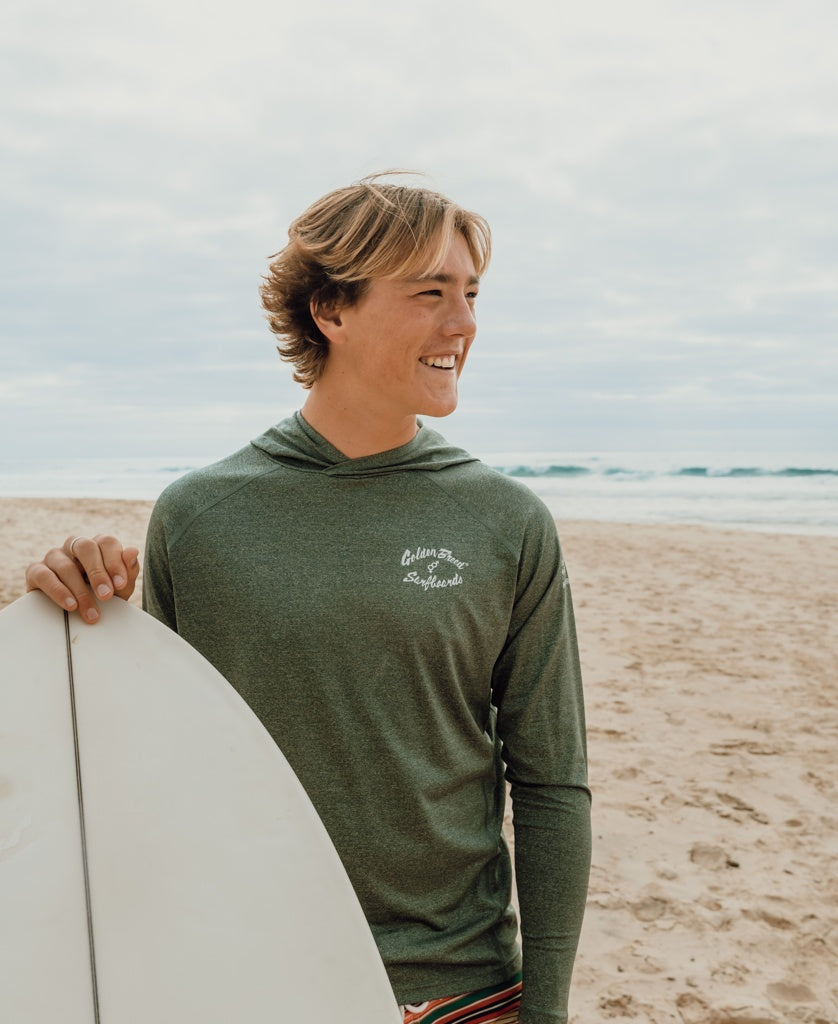 A young man stands on a sandy beach, smiling as he looks away, wearing the Golden Breed Rapper Hoodie Rash Shirt in Green Marle with the ocean in the background and holding a surfboard.