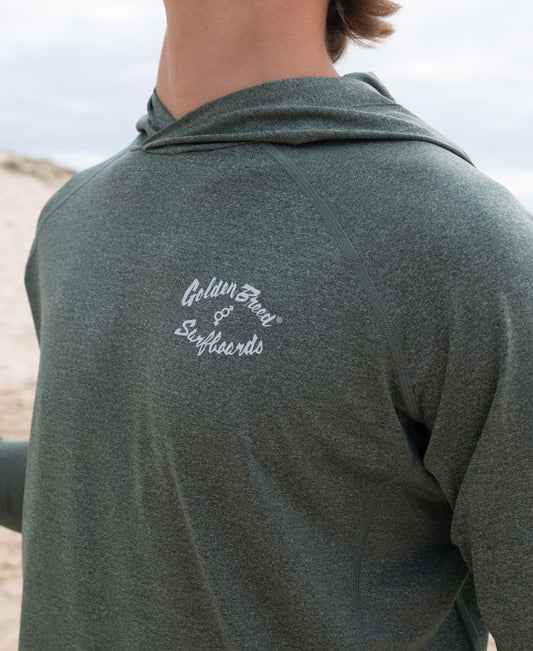 A person wears the Rapper Hoodie Rash Shirt in Green Marle by Golden Breed, featuring the logo on the chest, designed for surf enthusiasts, standing outdoors near sand dunes.