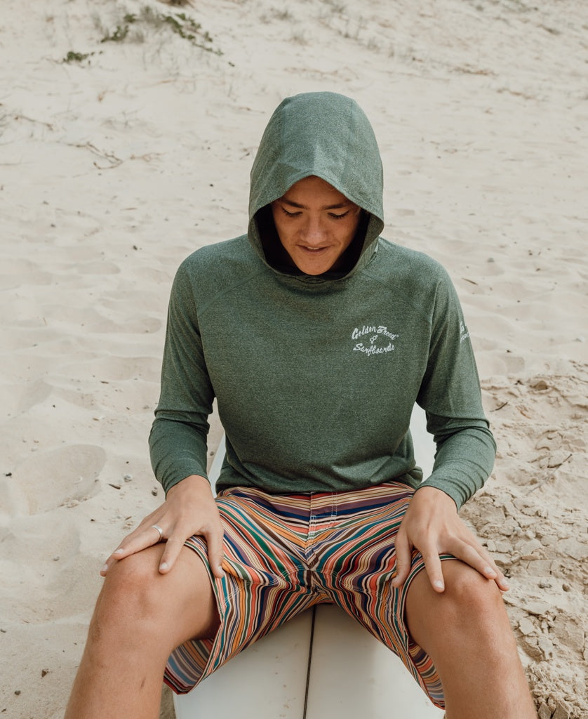 Wearing the Golden Breed Rapper Hoodie Rash Shirt in Green Marle and striped shorts, a person sits on a surfboard atop sandy dunes—looking down, ready to surf with 50+ UPF sun protection.