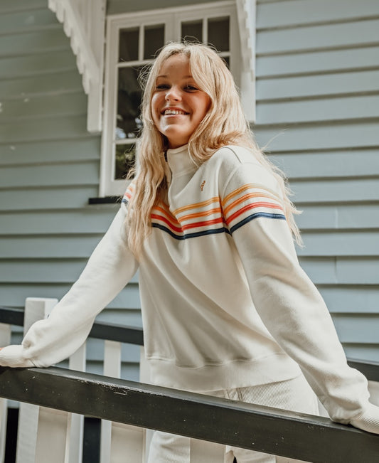 A smiling woman with long blonde hair wears the Golden Breed Long Island 1/4 Zip in off white, featuring colorful stripes and a retro logo, standing by a black railing in front of a light blue wooden house.