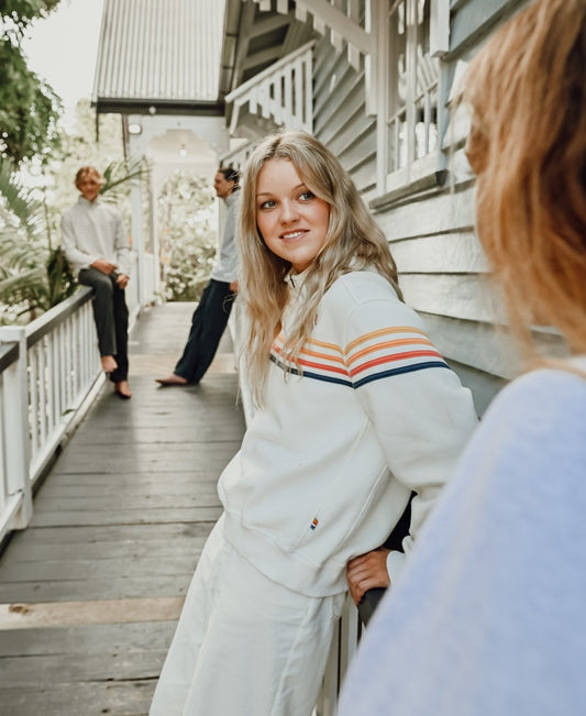 A young woman in the Golden Breed Long Island 1/4 Zip in Off White leans on a porch railing, smiling at someone, as two others chat near a grey wooden house.