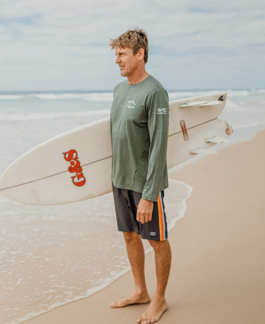 A man stands barefoot on the beach, wearing a Golden Breed Pebbles L/S Rash Shirt in Green Marle with 50+ UPF sun protection, holding a white surfboard under his arm and facing the ocean beneath overcast skies.