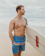 A shirtless, tattooed man stands on a beach with a surfboard, wearing Golden Breed’s Ralph Elastic Waist Boardie in Dusty Blue—crafted from recycled polyester—and gazes toward the ocean.