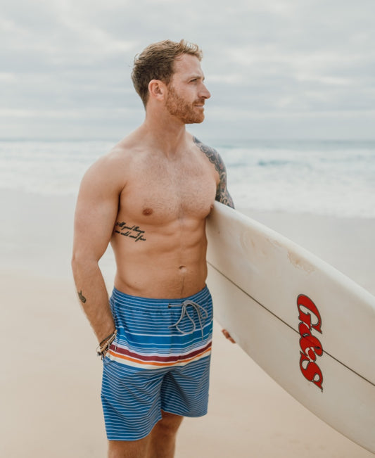 A shirtless, tattooed man stands on a beach with a surfboard, wearing Golden Breed’s Ralph Elastic Waist Boardie in Dusty Blue—crafted from recycled polyester—and gazes toward the ocean.