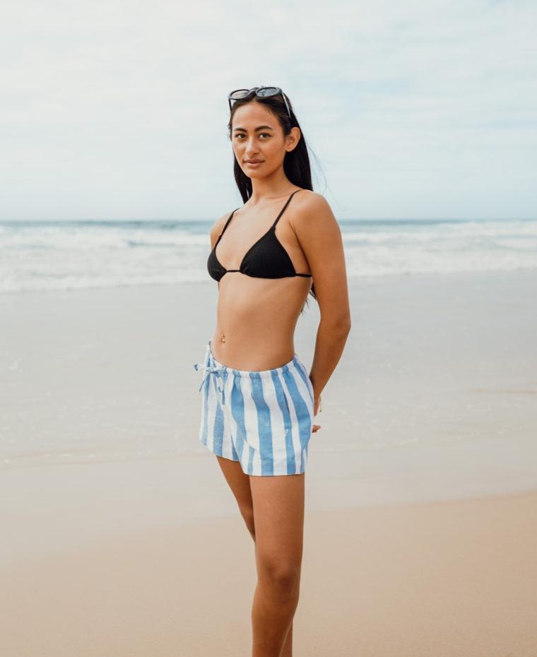 A woman wearing Golden Breeds Molly Short in Azure/White and a black bikini top stands on a sandy beach with the ocean in the background.