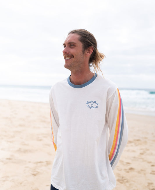 A man with long hair in a ponytail smiles on a sandy beach, wearing the Golden Breed Skater LS Tee in Off White. Colorful stripes accent the sleeves, and the ocean is visible behind him.