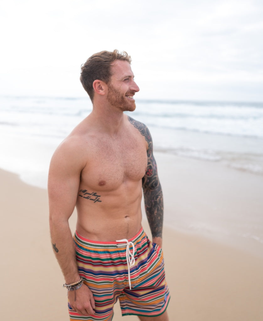 A bearded, tattooed man smiles on a sandy beach, wearing Golden Breeds Daze Elastic Waist Boardie | Stripe—swim trunks made from recycled polyester/spandex with 4-way stretch.