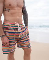 A man stands on a sandy beach with one hand in his pocket, wearing Golden Breeds Daze Elastic Waist Boardie | Stripe swim trunks; the ocean is visible in the background.