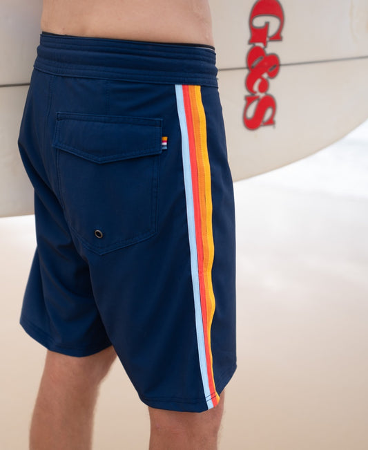 Wearing Golden Breeds Long Island Boardie in navy with a vibrant striped side, a person stands on the beach holding a white surfboard marked with red G&S letters, perfectly channeling the classic California surf vibe.
