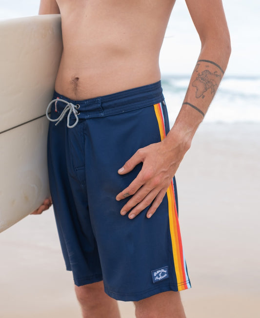 Wearing Golden Breeds Long Island Boardie in Navy, a person stands on the beach holding a surfboard, radiating California surf vibes—with part of an arm tattoo visible.