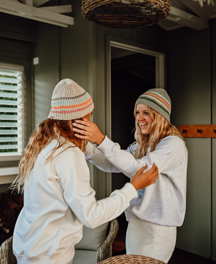 Two women smiling indoors wearing Organic Cotton Beanie Thin Stripe | Oatmeal in different stripe color variations, casual cozy style in a warm, retro setting.