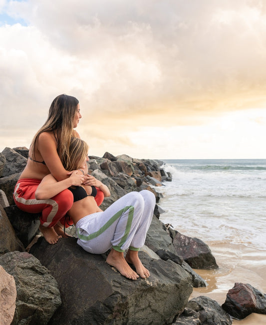 Two women sit on rocks by the beach at sunset, one hugging the other from behind as they gaze at the waves, both cozy in Golden Breedโs Retro Crunchie T/P | White Misty/Moss with signature stripes.