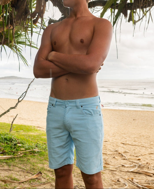 A shirtless person stands on a sandy beach with arms crossed, wearing Golden Breed Floyd Cord Walkshorts in Sky. The ocean and surfers can be seen in the background.