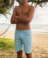 A shirtless person stands on a sandy beach with arms crossed, wearing Golden Breed Floyd Cord Walkshorts in Sky. The ocean and surfers can be seen in the background.