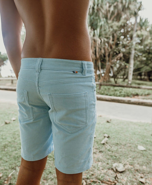 A person stands outdoors in light blue Floyd Cord Walkshorts by Golden Breed, seen from the back with grass and trees in the background.