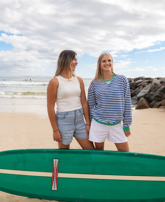 Two women stand on a sandy beach beside a green surfboard, both wearing the Ronnie Rib Tank in Off White by Golden Breed, with the ocean and rocks in the background under a partly cloudy sky.