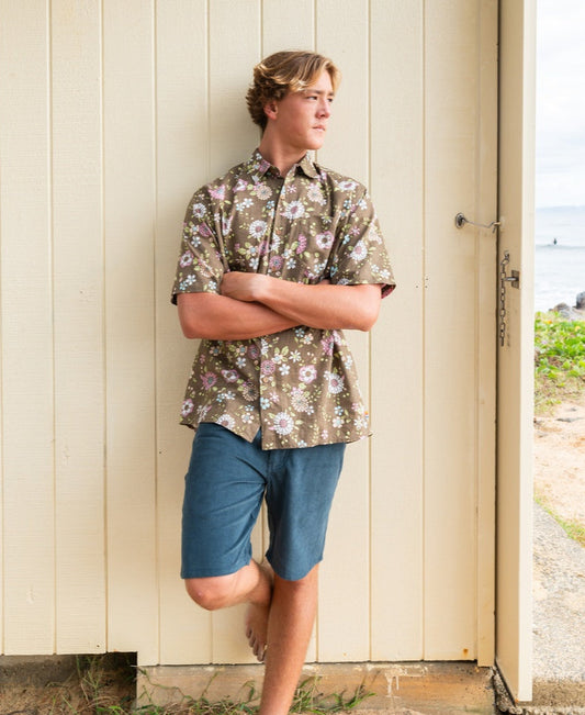 A young man stands with arms crossed, leaning against a light-colored wall near a beach, wearing the Golden Breed Happy Floral Shirt in Wash Brown and blue shorts.