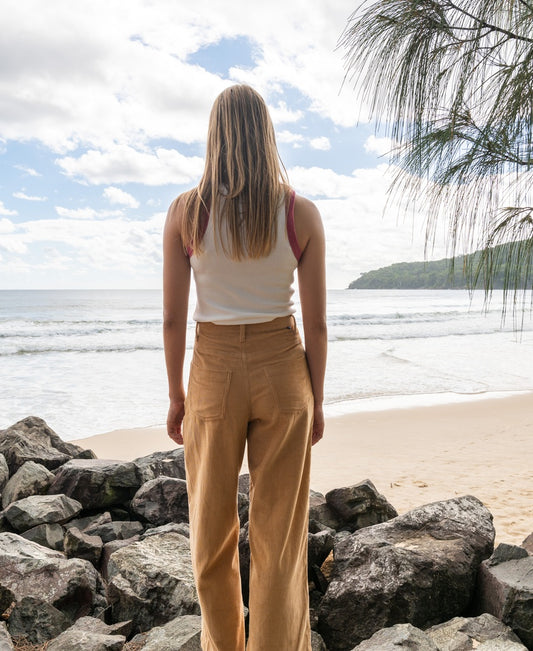 Wearing a white tank top and Golden Breeds Jenny Cord Pants in Camel, a long-haired person stands on rocks facing the ocean on a partly cloudy day.