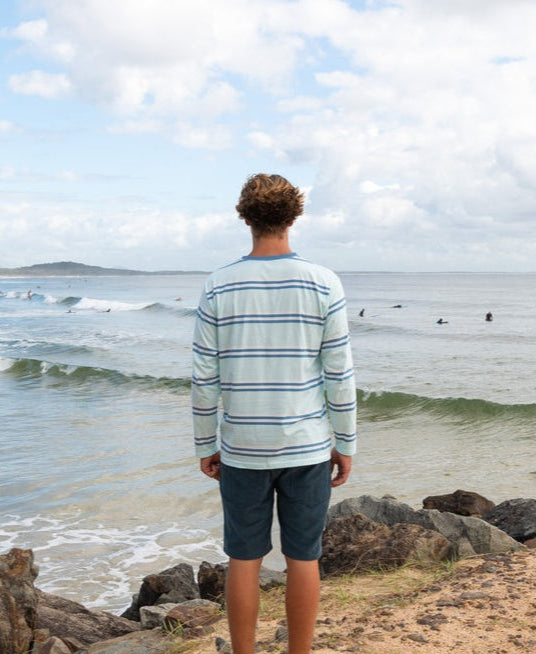 Wearing the Golden Breed Bay Stripe LS Tee | Ice and shorts, a person stands on rocks facing the ocean, soaking up the Cali surf vibe as surfers catch waves beneath a cloudy sky.