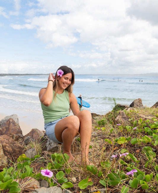 A woman sits on seaside rocks, smiling as she tucks a pink flower in her hair. She wears the Golden Breed Ronnie Rib Tank in Moss and denim shorts, with surfers and the ocean in the background. The tank is handmade in Indonesia.