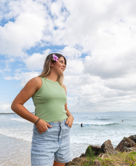 A woman in the Golden Breed Ronnie Rib Tank | Moss and denim shorts stands by the ocean with a flower in her hair, hands in pockets. The handmade-in-Indonesia tank is paired with a cloudy sky and surfers seen offshore.