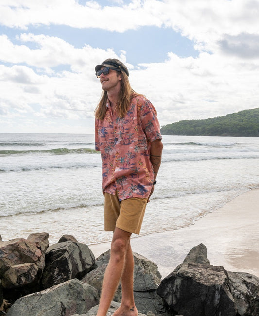 Wearing the Flamingos Shirt | Rose by Golden Breed, tan shorts, sunglasses, and a cap, a person stands barefoot on beach rocks with waves and a forested hill in the background.
