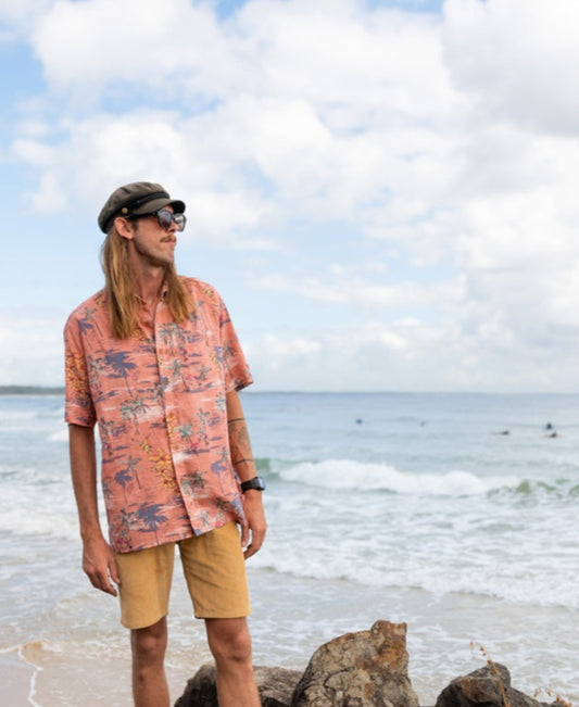 A man wearing the Golden Breed Flamingos Shirt | Rose, with shorts, sunglasses, and a hat, stands on rocks by the ocean as waves crash under a cloudy sky.
