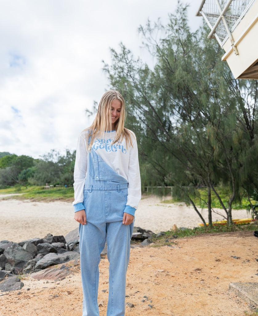 A person with long blonde hair wears the Maci Overall in Light Blue by Golden Breed over a white sweatshirt, standing on sand near rocks and trees, with a beach and cloudy sky in the background.