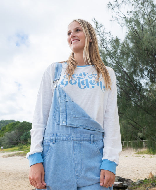 A young woman smiles outdoors in light blue denim overalls layered over the Golden Breed Stay Golden LS | Off White/Azure—a 100% cotton, cropped long sleeve tee with blue cuffs and golden text. Trees and sand create a natural backdrop.