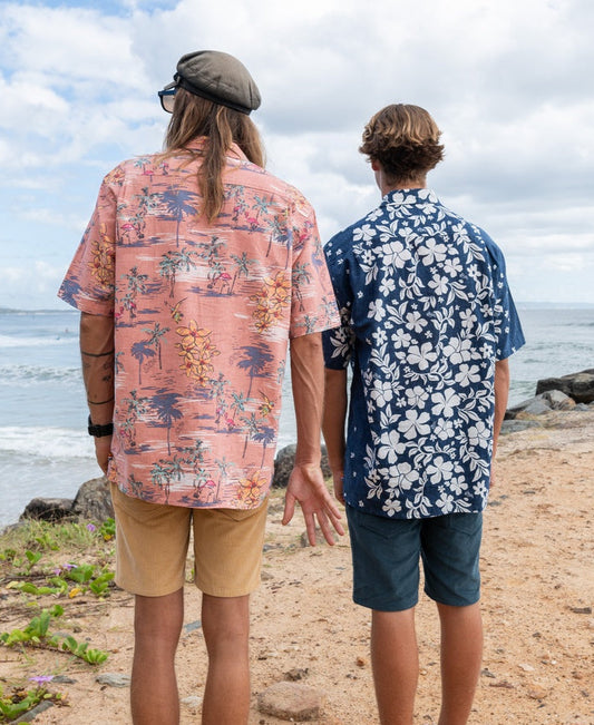 Two people stand side by side on sandy ground near rocks and greenery, facing the ocean while wearing Golden Breedโs Vomo Floral Shirt in Vintage Navy.