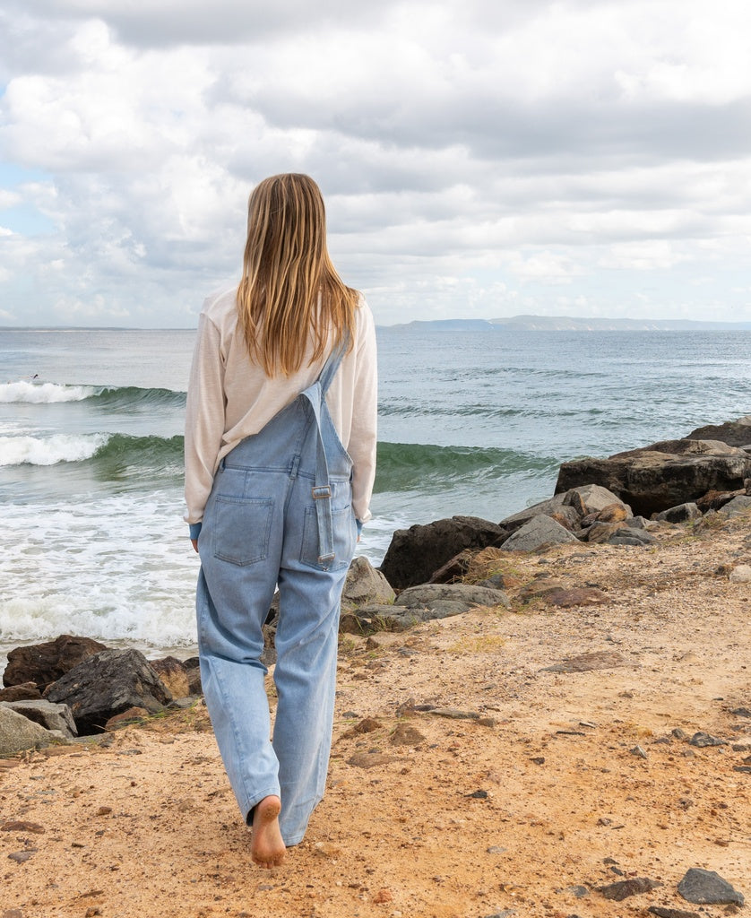 A person with long hair, barefoot and facing away, walks on a sandy path by the ocean near rocks, wearing a white shirt and Golden Breeds Maci Overall in light blue with adjustable straps.
