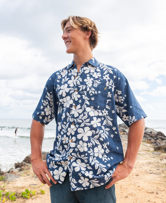 A young man stands on a beach smiling and looking aside, wearing the Golden Breed Vomo Floral Shirt in Vintage Navy.