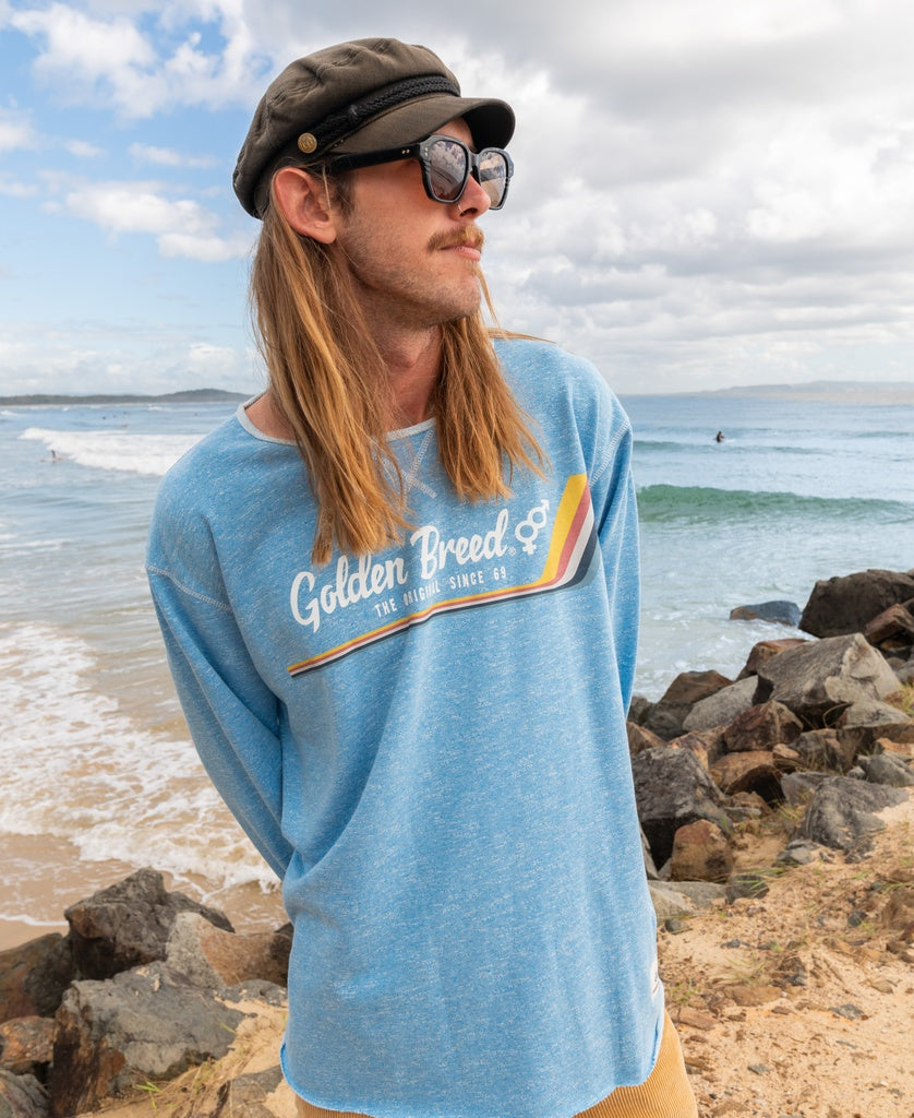 A man with long hair, cap, and sunglasses wears a Golden Breed Retro Rag Top in Sky Marle on a rocky beach with the ocean behind him.