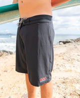 Wearing Gordon & Smiths G&S Boardie in Black, made from recycled polyester, a person stands on a sandy beach near the ocean with waves and rocks in the background.