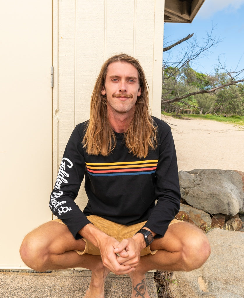 A man with long hair wearing the Golden Breed Heavy Island Long Sleeve Tee in black and tan shorts squats outdoors by a pale wall and rocks, channeling a chill Cali surf vibe.