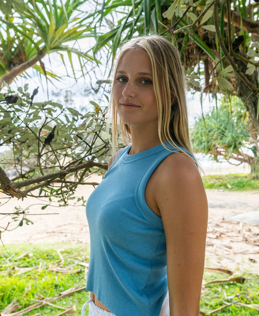 A young woman wears the Golden Breed Ronnie Rib Tank in Azure, standing outdoors by green foliage and gazing at the camera with a neutral expression.