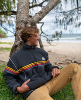 Wearing a Golden Breed Reef Bonded Sherpa Jacket in Wash Black, a person sits on the grass by a tree, looking at the sandy beach and ocean under cloudy skies. The cozy polyester sherpa provides warmth in this serene moment.