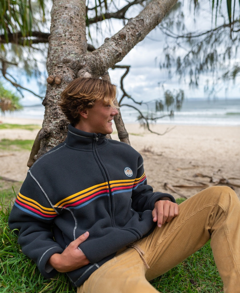 Wearing a Golden Breed Reef Bonded Sherpa Jacket in Wash Black, a person sits on the grass by a tree, looking at the sandy beach and ocean under cloudy skies. The cozy polyester sherpa provides warmth in this serene moment.