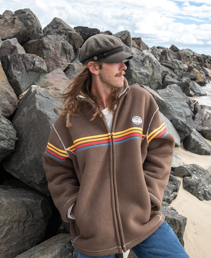 A man with long hair wearing a cap and a Golden Breed Reef Bonded Sherpa Jacket in Wash Brown with colorful stripes stands by large rocks on the beach, gazing to the side.