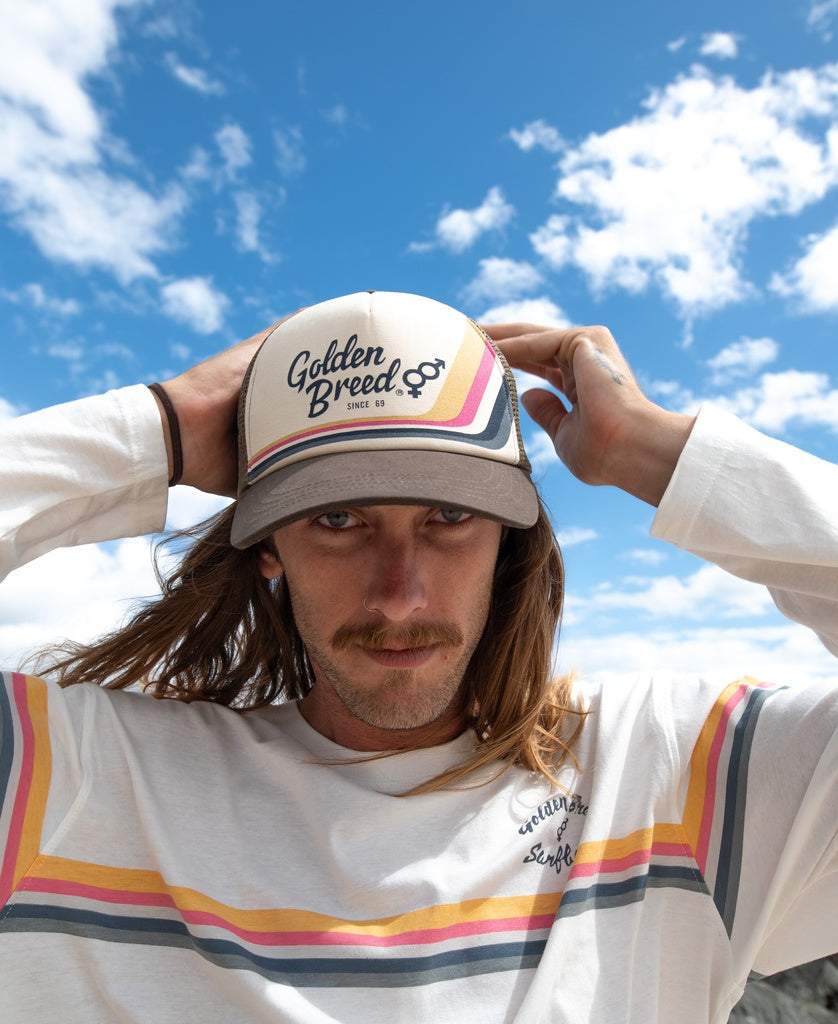 A person wearing a Golden Breed Retro Trucker Cap in Wash Brown with an adjustable strap and a matching striped shirt stands outdoors under a blue sky with scattered clouds.