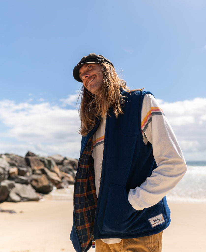 A person with long hair, wearing a Golden Breed Thredbo Vest in Midnight, a striped long-sleeve shirt, and a cap, stands on a sandy beach near rocks under a partly cloudy sky.