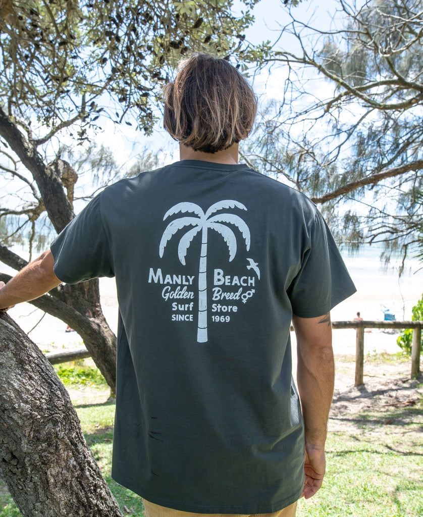 Facing away outdoors with trees and a beach in the background, a person wears the Golden Breed Manly Palm Loco Tee in Wash Black, showcasing a white palm tree graphic and Manly Beach Golden Breed Surf Store Since 1969.